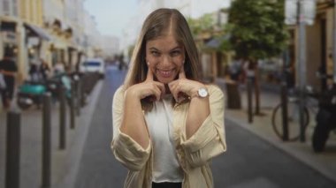 Genç sarışın kadın, bulanık vitrinler ve park etmiş scooterlarla dolu kalabalık bir sokakta parmaklarıyla neşe içinde yanakları itiyor. Mutluluk..