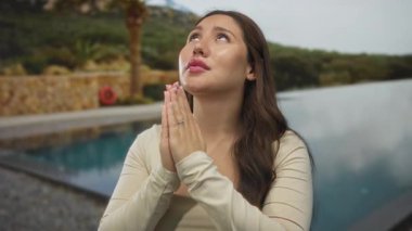 Elleri duada birbirine bastırılmış genç İspanyol bir kadın bir tatil köyünde açık havuz kenarına bakıyor..
