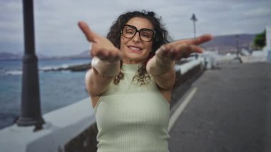 Kıvırcık saçlı, kolsuz üst ve siyah çerçeveli gözlüklü bir kadın gülümser ve deniz kenarında kollarını uzatır..