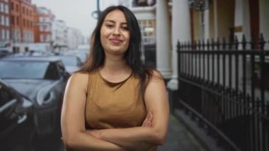 Kolları çapraz ve eğimli bir kadın, şehir caddesinde kahverengi kolsuz gömlek giyiyor; özgüven özgünlüğü tavrı.