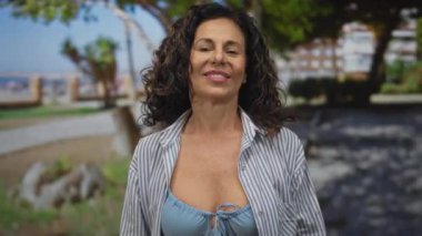 Parkta elleri bikini ve çizgili gömlekle gülümseyen ve el kol hareketi yapan bir kadın. Açık havada güneşli bir günün tadını çıkarıyor..