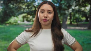 Beyaz gömlekli genç İspanyol kadın yeşil ormanda kollarını kavuşturuyor ve doğal ışık altında ciddi bir ifade sergiliyor..