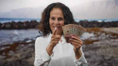 Elinde Kolombiya pezosu olan bir kadın, deniz kenarında gülümsüyor, kumsalda banknotlar ve arka planda dalgalar sergiliyor..