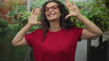 Kırmızı gözlüklü bir kadın iki eliyle yüzünü çerçeveliyor ve ormanda gülümsüyor. Neşeli bir sevinç..