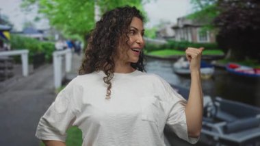 Beyaz tişörtlü kadın başparmağıyla kalabalık şehir sokaklarında kanalı işaret ederek gülümsüyor; seyahat heyecanı.