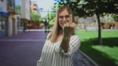 Etrafında bulanık binalar ve kaldırımlarla çevrili güneşli şehir sokağında kameraya doğru orta parmak hareketi yapan bir kadın..