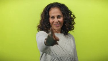 Kıvırcık saçlı ve kameraya göz kırpan olgun bir kadın, izole edilmiş sarı bir arka plana yaslanmış, neşe ve kendine güveni ifade ediyor..