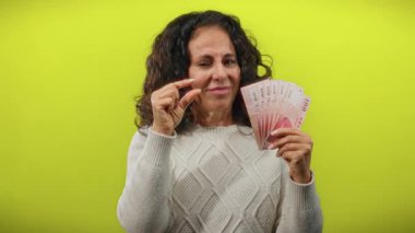 Tayvanlı orta yaşlı bir kadın, izole edilmiş sarı arka plan duvarına dolar banknotları tutarken küçük beden jestler yapıyor..