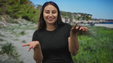 Gün ışığında kameraya bakarken kumlu bir sahil sahilinde açık avuçlarla gülümseyen kadın; sıcak davetkar neşe.