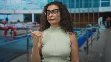 Kıvırcık saçlı ve gözlüklü genç İspanyol kadın, gemi güvertesinde tırabzan döşeyerek baş parmağını yana gösteriyor..