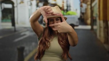 Uzun saçlı genç İspanyol kadın, gündüz vakti bir caddede gülümseyen elleriyle yüzünü çerçeveler..