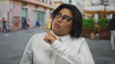 Gözlüklü, iri yarı Latin bir kadın canlı bir şehir caddesinde düşünceli bir şekilde el kol hareketi yapıyor, açık havada çeşitlilik ve güven gösteriyor..