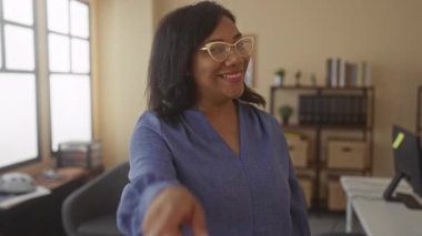 Ofis binasında kamerayı işaret eden bir kadın, gözlük ve mavi bluz takıyor ve dosya ve kutuların arasında duruyor..