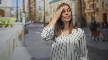 Çizgili tişörtlü genç sarışın kadın gün ışığı altında bina dışında elle alnını siliyor..