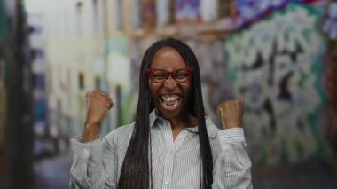 Örgü örgülü ve gözlüklü bir kadın sokak ortasında gülümsüyor graffiti arka planında şehir ortamında neşe ve tarz sergiliyor..