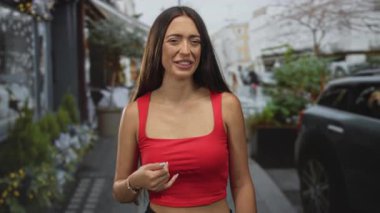 Kadın şehir caddesinde işaret işareti işareti yapıyor, kırmızı ekin üst omuz ve göbeğini gösterirken parmak ucunu gösteriyor; davetiye kendinden emin.