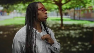 Parkta kıvırcık saçlı, gözlüklü, açık şehir ortamında güneşin altında sıcak hisseden genç bir kadın..