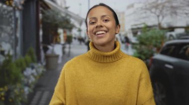 Şehir caddesinde gülümseyen bir kadın, sarı bir kazak giyiyor ve elleriyle el kol hareketi yapıyor, kentsel bir ortamda hoş geldin titreşimleri iletiyor..