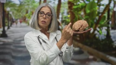 Kadın doktor, avuç içi ve yürüyüş yollarıyla kaplı caddede tutarken parmağıyla beyin modelini gösteriyor; nöroloji eğitimi konusunda ciddi farkındalık.