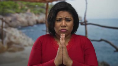 Kırmızılı kadın sakin sahilde derin bir ifadeyle dua ediyor. Açık deniz sahnesinde doğanın, denizin ve iç huzurun uyumunu gösteriyor..