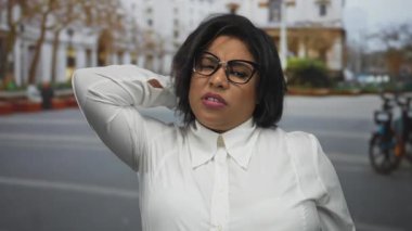 Şehir caddesinde boynu ağrıyan bir kadın, elleri boynunda, beyaz gömlek ve gözlük takmış, bulanık binaları ve bisikletleri olan bir şehir zeminine karşı rahatsızlığını dile getiriyor..