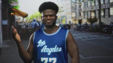 Genç Afrikalı Amerikalı adam mavi basketbol forması ve geniş bir gülümseme içinde meşgul şehir caddesinde el çırpıyor; sevinç kutlaması.