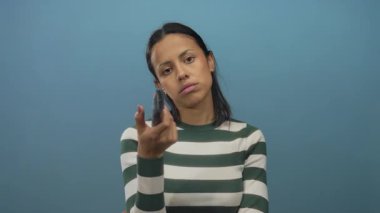 Televizyonun karşısında uzaktan kumandayı elinde tutan genç bir kadın, izole edilmiş mavi bir arka planda, odaklanmış ve düşünceli görünüyor..