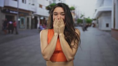 Turuncu ekin giymiş bir kadın, bulanık vitrinleri olan bir yaya caddesinde şaşkınlıkla el ele tutuşuyor..