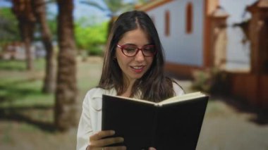 Kırmızı gözlüklü İspanyol kadın yeşil orman ortamında parmak işareti yapıyor; davetiye.