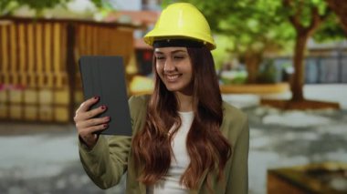 Sert şapkalı İspanyol kadın mühendis parkta neşeli bir video görüşmesi yapıyor, profesyonel bağlantı ve teknoloji kullanımını doğal ortamında gösteriyor..