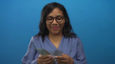 Mavi stüdyoda para sayan, gözlüklü ve gülümseyen bir kadın var..