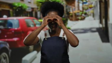 Önlüklü kadın, arka planda arabaları ve binaları olan bir şehir caddesinde dürbünlü jestler yapıyor..