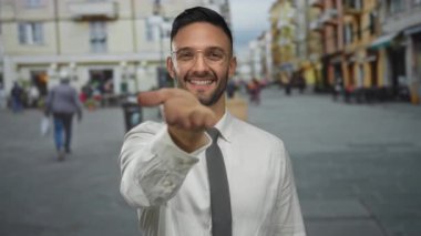 Meşgul bir şehir sokağında gülümseyen gözlüklü genç İspanyol adam, arka planda insanlarla birlikte canlı bir şehir manzarasında elini uzatarak davetkar bir jest sergiliyor..