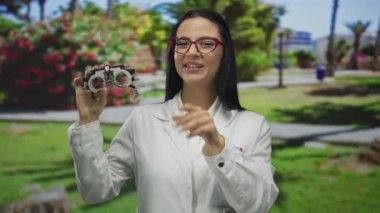 Optisyen kadın dışarıda güneşli bir parkta optik ekipman tutuyor yemyeşil ve parlak çiçekli, görüş alanında profesyonel uzmanlık sergiliyor.
