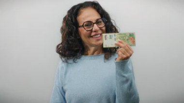 Gülümseyen kadın, orta yaş ve finansal kavramları vurgulayan beyaz bir arka plan karşısında günlük süveter içinde Arjantin pesoları tutuyor..