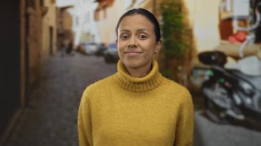 Rahat sarı bir süveter içinde gülümseyen bir kadın güvenli bir şekilde bir şehir sokağında duruyor, huzur ve sıcaklığı somutlaştırıyor kırsal bir ara sokakta..