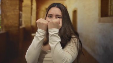 Eski bir üniversite binası koridorunda uzun kollu bluz giyen, yumruklarını kaldırmış ve gergin yüzlü bir kadın. Kararlılık, kendine güven dayanıklılığı..