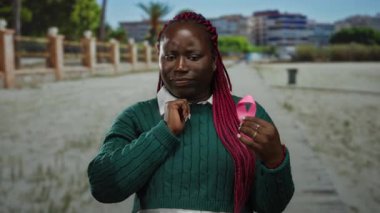 Yeşil kazaklı kadın parkta pembe kurdele tutuyor. Arka planda binalar var. Açık hava ortamında meme kanseri farkındalığı ve desteğine odaklanıyor..