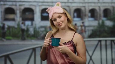 Gece elbiseli bir kadın Roma 'da kolezyum arka planında bir kupa tutuyor, neşeli bir şekilde gülümsüyor, arka planda ikonik italyan mimarisi ile rahatlamış sabah titreşimlerini somutlaştırıyor..