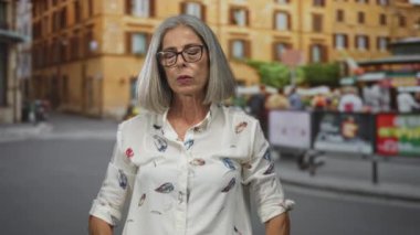 Kolları açık ve gözlüklü kadın şehir caddesinde, gözleri kapalı ve yukarı bakıyor; dinginlik yansıması.