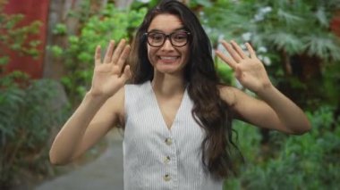 Çizgili üst dalgalar takan kadın yeşil parktaki kameraya doğru elini uzatıyor. Bulanık yaprakları ve görülebilir bir ağaç gövdesi var..