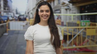 Sokak inşaatı bariyerleri ve sarı güvenlik rayları yanında beyaz gömlekli, görünür dişleriyle göz kırpan ve gülümseyen genç İspanyol kadın..