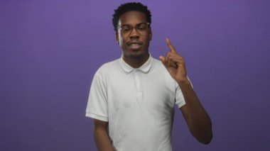 Gözlüklü ve beyaz polo tişörtlü bir adam mor stüdyoda hafif ışıklandırmanın altında parmağını yukarı kaldırıyor..