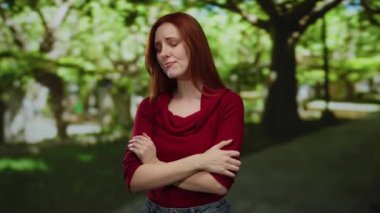 Güneşli bir parkta kolları çapraz duran bir kadın, sakin bir günde yemyeşil ağaçların arasında merak ve tefekkür ifadesi sergiliyor..