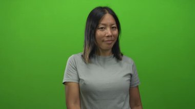 Gri tişörtlü kadın elinde kamerayla stüdyoya doğru parlak yeşil duvarlı, hoş geldin jesti.