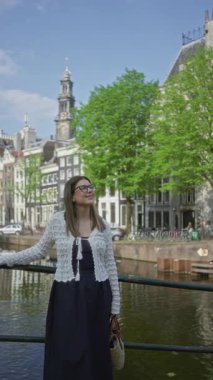 Amsterdam 'da güneşli bir günde arka planda tarihi binalar ve mavi gökyüzünün altında mutlu bir şekilde gülümseyen hasır çanta tutan bir kadın..