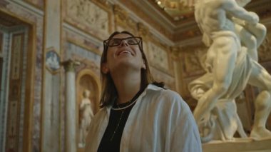Gözlüklü kadın, klasik heykellerle çevrili dekoratif müze binasında yüzü görünen mermer heykele bakıyor..