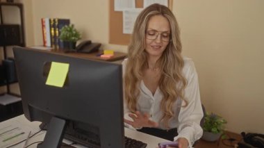 Kadın ofiste bilgisayarla oturup gülümserken selfie çekiyor, neşeli ve modern bir çalışma alanı ortamı yaratıyor..