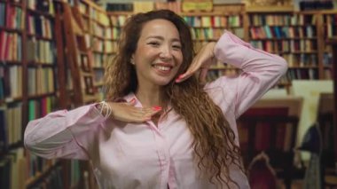 Kütüphanede kırmızı tırnaklar ve pembe gömlek gösteren genç bir kadın gülümsüyor ve elleriyle yüzünü çerçeveliyor; neşeli öğrenme.