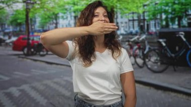 Kadın bir şehir caddesinde eli ile ağız kaplayan beyaz t-shirt giyen ve kalkan yüz gülümseyen; oyuncu sessizlik.